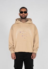Beige hoodie med en kænguru lomme og grafisk tekst. Har lilla sommerfugle detaljer på brystet. Bæres med sorte cargo bukser og solbriller.