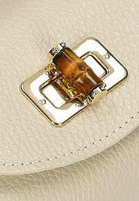 Cremefarbene Ledertasche mit einer goldenen rechteckigen Schnalle und einem zentralen Holzakzent, die eine strukturierte Oberfläche und Detailnähte hervorhebt.