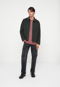 Mørkegrønn zip-up jakke med krage, to frontlommer, båret over en mauve t-skjorte, kombinert med mørke cargo-bukser og svarte sko.