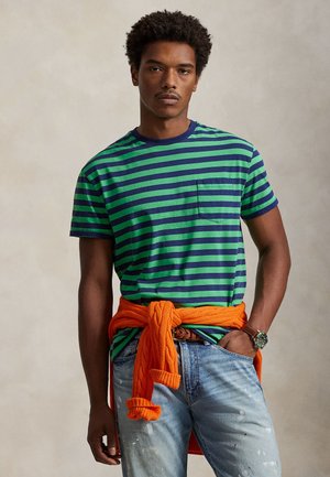 Ριγέ μπλουζάκι με κοντά μανίκια σε πράσινο και ναυτικό μπλε, με τσέπη. Φορεμένο με ξεθωριασμένο μπλε τζιν και πορτοκαλί πουλόβερ δεμένο στη μέση.