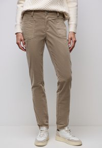 Persona con pantalones beige de pierna recta y zapatillas blancas, de pie contra un fondo gris claro liso.