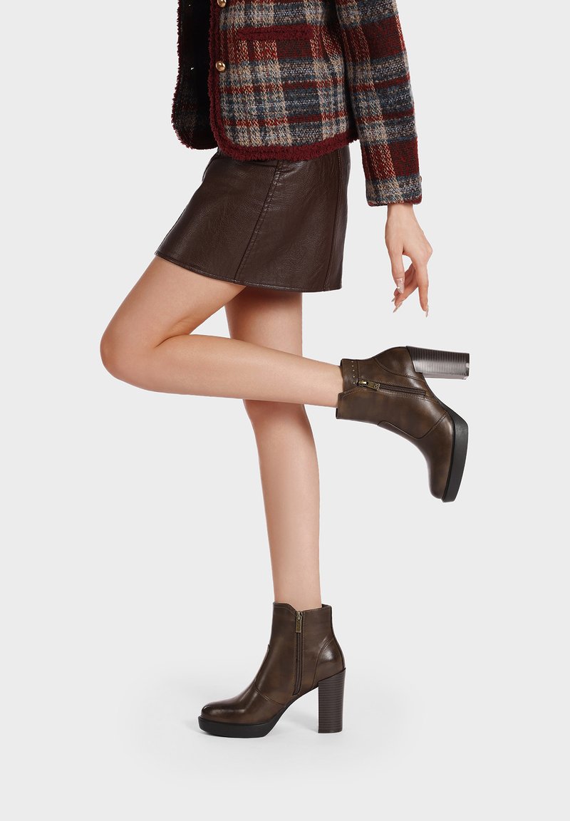 Donna che indossa stivaletti in pelle marrone con tacco spesso, una mini gonna in pelle marrone scuro e una giacca di lana scozzese, con una gamba sollevata all'indietro.