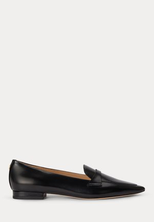 Zapato plano de cuero negro con punta afilada, tacón bajo y detalle de correa en la parte delantera, mostrado en perfil lateral sobre un fondo blanco.