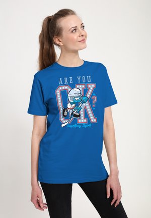 Jeune femme portant un T-shirt bleu avec un Schtroumpf jouant au hockey et le texte « Ça va ? Esprit Schtroumpf. »