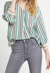 Blusa a righe verdi e bianche con scollo a V, maniche lunghe e polsini con dettagli dorati; abbinata a jeans grigi attillati.