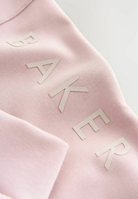 Lichtroze sweatshirtstof met verhoogde witte letters die "BAKER" spellen, met een gladde textuur en een licht gebogen ontwerp.