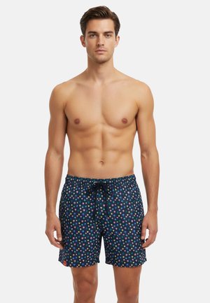 Giovane uomo in piedi, senza maglietta, che indossa pantaloncini da bagno blu navy con un piccolo motivo floreale colorato e vita con coulisse.