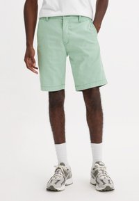 Uomo che indossa pantaloni corti verde chiaro al ginocchio, calze bianche e sneakers bianco-grigio, in piedi con una mano in tasca.