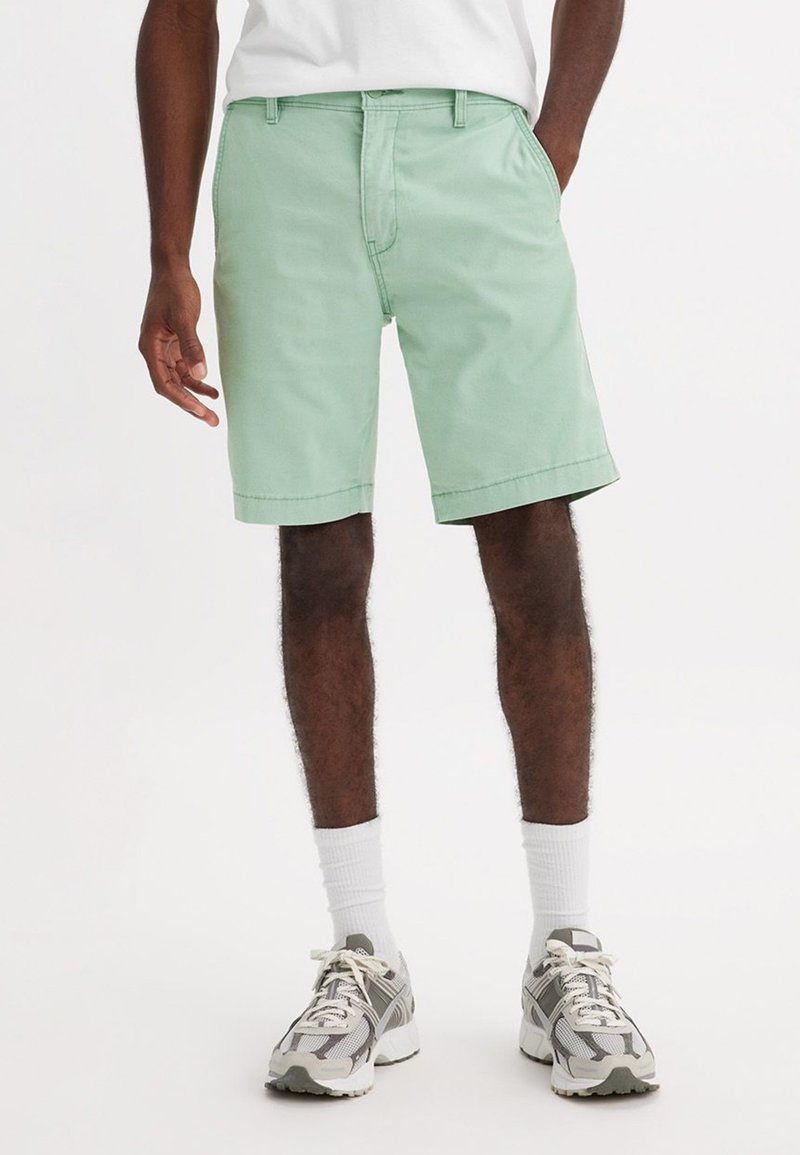 Uomo che indossa pantaloni corti verde chiaro al ginocchio, calze bianche e sneakers bianco-grigio, in piedi con una mano in tasca.