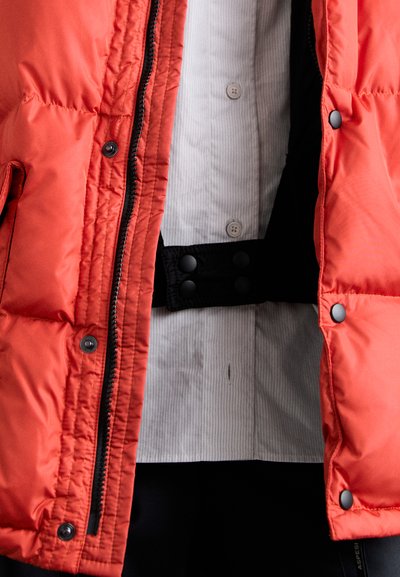 Rode nylon pufferjack met ritsluiting, drukknoopjes en een zwarte riem. Een wit gestreept shirt is zichtbaar eronder, wat een textuurcontrast toevoegt.