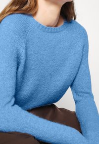 Maglione blu in lana con scollatura rotonda e maniche lunghe. Texture morbida e vestibilità comoda, abbinato a un indumento marrone sottostante.