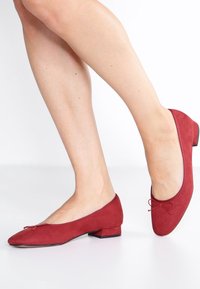 Les ballerines en daim rouge présentent un bout rond, un petit talon bloc et un nœud décoratif à l'avant. Le design est élégant et minimaliste.