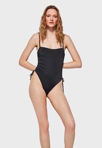 Bañador negro de una pieza con tirantes finos ajustables, detalles fruncidos en los laterales, abertura de pierna alta y lazos laterales para un ajuste personalizable.