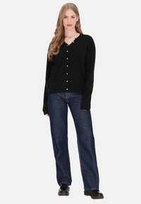 Fekete, V-nyakú kardigán gyöngyszerű gombokkal az elején, kék bő szárú farmerrel és fekete cipővel párosítva. Puha anyagú.