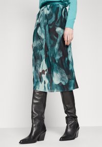 Jupe midi plissée en imprimé abstrait teal et noir, fabriquée en tissu texturé, associée à des bottes noires en cuir avec talons, arrivant au genou.