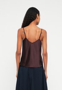 Kobieta z długimi kręconymi włosami, ubrana w ciemnofioletowy top bez rękawów na cienkich ramiączkach oraz granatową spódnicę, pokazana od tyłu na białym tle.