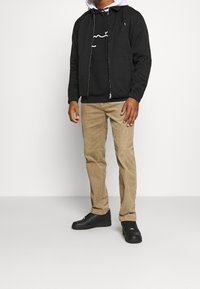 Giacca nera con coulisse, felpa bianca sotto, design grafico nero sulla felpa, pantaloni in corduroy beige e sneakers nere.