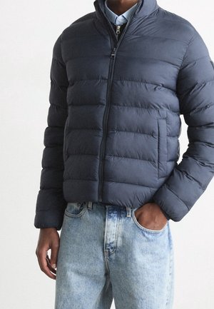 Homme portant une veste matelassée bleu marine zippée par-dessus une chemise à col bleu clair avec un jean bleu délavé clair.