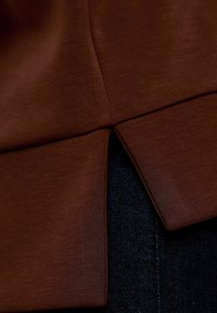 Top in tessuto marrone con superficie liscia e spacco laterale, posizionato su denim scuro con cuciture visibili e una leggera lucentezza.