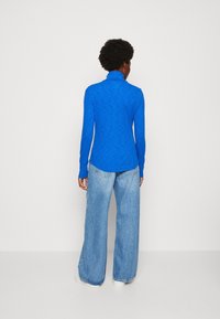 Top blu lungo con maniche, colletto alto e tessuto texture, abbinato a jeans larghi blu chiaro e sneaker bianchi, visto da dietro.
