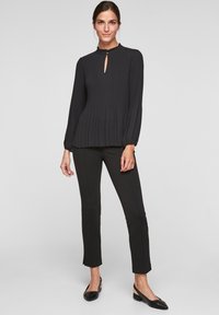 Zwarte geplooide blouse met een keyhole halslijn, lange mouwen en een licht uitlopende zoom, gecombineerd met strakke zwarte broek en platte schoenen.