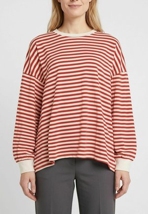Vrouw draagt een rood-wit horizontaal gestreept shirt met lange mouwen en witte manchetten, en donkergrijze broek, kijkt recht vooruit.