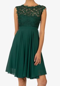 Grön klänning med spetsliv, ärmlös design, plisséad kjol och åtsittande midja. Spetsen har ett blommönster och genomskinliga detaljer.