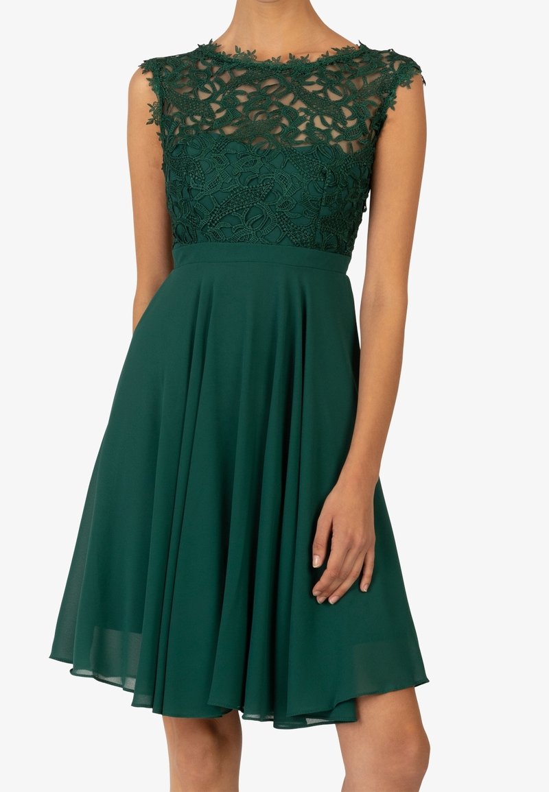 Grön klänning med spetsliv, ärmlös design, plisséad kjol och åtsittande midja. Spetsen har ett blommönster och genomskinliga detaljer.