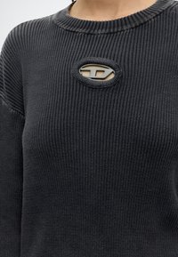 Gros plan d'une personne portant un pull noir côtelé avec une petite découpe ovale révélant un motif beige et noir en dessous.