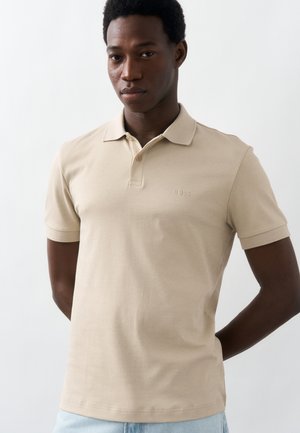 Νεαρός άνδρας που στέκεται με τα χέρια πίσω από την πλάτη, φοράει μπεζ κοντομάνικο πόλο και ανοιχτό μπλε τζιν, με φόντο ένα απλό λευκό τοίχο.