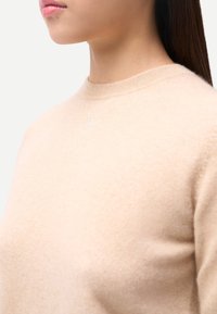 Pull beige clair avec un col rond et une texture subtile. Présente un logo brodé au centre. Tissu doux et confortable.