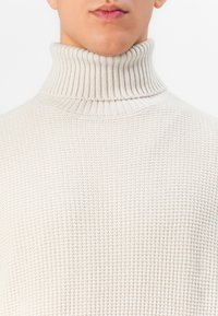 Close-up of a person wearing a cream-colored ribbed turtleneck sweater with a textured knit pattern.