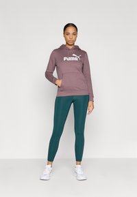 Burgundská mikina s bielym logom Puma, predná vrecko a kapucňa na šnúrku; skombinovaná s azúrovými legínami; biele tenisky dopĺňajú outfit.