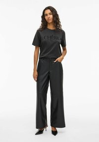 Sort T-shirt med teksten "CHÉRIE", parret med brede sorte læderbukser. Glat tekstur; modellen bærer sorte højhælede sko.