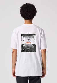 White cotton t-shirt featuring a graphic print on the back with concentric circular patterns in black and orange accents. Short sleeves.