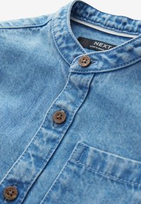 Camisa de mezclilla en azul claro con cuello redondo, tres botones de madera y un solo bolsillo en el pecho. Textura suave con áreas desgastadas.