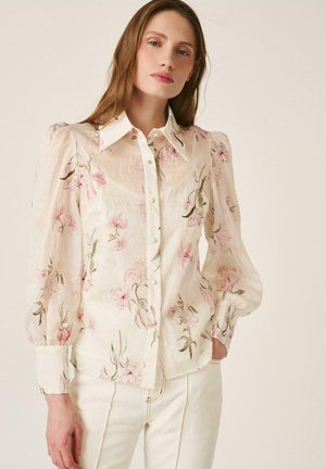 Mujer con una blusa blanca transparente con estampados florales rosas y mangas abullonadas, combinada con pantalones blancos de talle alto, de pie con una mano en el bolsillo.