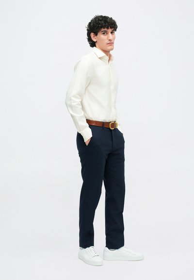 Slim-fit bílá košile, tmavě modré kalhoty, hnědý kožený opasek se zlatou sponou a bílé tenisky. Model stojí s rukama v kapsách.