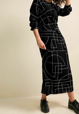 Femme portant une robe noire à manches longues avec des lignes géométriques blanches et des chaussures noires à lacets, debout devant un fond beige uni.