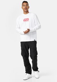 Valkoinen pitkähihainen t-paita, jossa on punainen "Star Wars: The Empire Strikes Back" -grafiikka, yhdistettynä mustiin cargo-housuihin ja valkoisiin lenkkareihin.