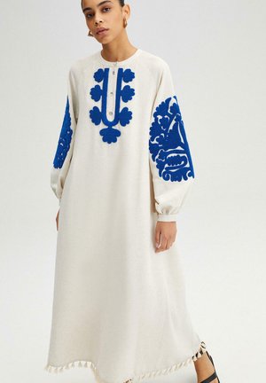 Femme portant une longue robe crème avec une broderie florale bleue sur les manches et la poitrine, se tenant devant un fond clair uni.