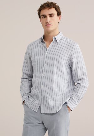 Junger Mann, der ein hellgraues Hemd mit vertikalen Streifen und eine hellgraue Hose trägt, steht mit den Händen in den Taschen vor einem beigen Hintergrund.