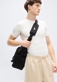 Black crossbody bag with adjustable strap, fabric texture, and multiple compartments, worn over a fitted white shirt and beige sweatpants.
