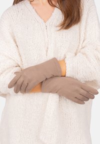 Braune Wildlederhandschuhe mit glatter Textur und schmal zulaufenden Fingern, getragen von einer Person mit einem cremefarbenen Strickpullover.