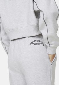 Szary zestaw dresowy, składający się z bluzy i spodni, z przeszywanymi detalami oraz haftowanym logo na spodniach. Miękka tkanina, luźny krój i kieszenie.