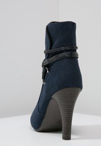 Marinblå mocka stövlett med en hög, avsmalnande klack, prydd med ett flätat svart band och en justerbar spännros. Slät yta över hela skon.