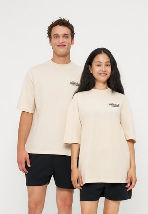 Junger Mann und Frau, die nebeneinander stehen und übergroße beige T-Shirts mit schwarzem Text sowie schwarze Shorts vor einem schlichten hellen Hintergrund tragen.