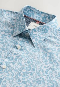 Camisa floral de color azul claro con un cuello y botones estampados. Presenta diseños intrincados de flores sobre un fondo blanco. Etiqueta visible dentro del cuello.