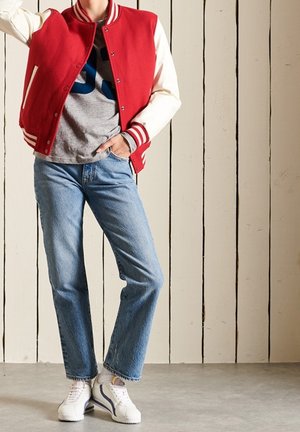Giacca varsity rossa e bianca, t-shirt grigia con grafica, jeans blu, sneakers bianche con dettagli viola; in piedi contro un muro di legno.