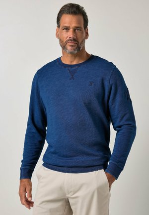 Hombre con barba que lleva un suéter azul de manga larga y pantalones beige claros, de pie contra un fondo claro y liso.